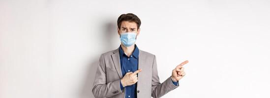 Covid-19, pandemic and business concept. Upset man in medical mask and office suit, frowning sad and pointing left at banner, standing on white background photo