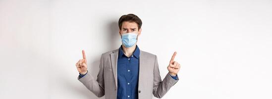 Covid-19, pandemic and business concept. Disappointed frowning guy in suit and medical mask pointing fingers up, complaining on unfair situation, white background photo