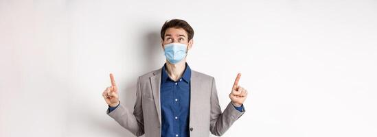 Covid-19, pandemic and business concept. Handsome businessman in medical mask and suit, looking and pointing up with dreamy face, white background photo
