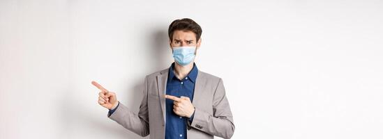 Covid-19, pandemic and business concept. Disappointed and reluctant man in medical mask frowning, pointing fingers right at something disgusting or ugly, white background photo