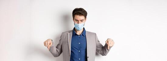 Covid-19, pandemic and business concept. Surprised guy in medical mask and suit looking and pointing down with popped eyes, gasping amazed, white background photo