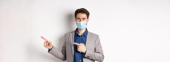 Covid-19, pandemic and business concept. Confident male manager in suit and medical mask showing way, pointing fingers right at advertisement, white background photo