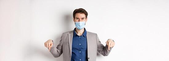 Covid-19, pandemic and business concept. Handsome businessman in medical mask and suit, pointing fingers down and showing company logo, white background photo