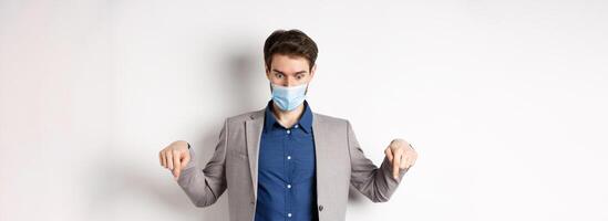 Covid-19, pandemic and business concept. Shocked guy in medical mask and office suit pointing, looking down startled, white background photo