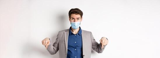 Covid-19, pandemic and business concept. Hesitant man in suit and medical mask asking opinion, pointing fingers down and look at camera, white background photo