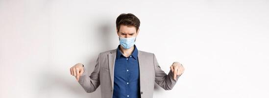 Covid-19, pandemic and business concept. Confused and displeased businessman in medical mask frowning, pointing fingers down and looking disappointed, white background photo