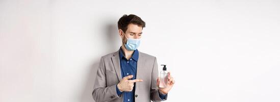 Covid-19, pandemic and business concept. Office worker in medical mask pointing at bottle of hand sanitizer, using antiseptic at work, white background photo