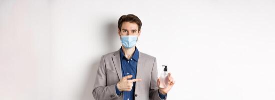 Covid-19, pandemic and business concept. Office worker in medical mask pointing at bottle of hand sanitizer, using antiseptic at work, white background photo