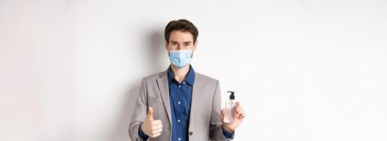 Covid-19, pandemic and business concept. Businessman in office suit and medical mask showing bottle of hand sanitizer and thumb up, recommend use antiseptic at work photo