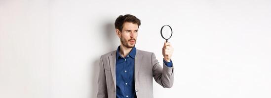 Serious man in suit searching for clues through magnifying glass, investigating, standing on white background photo
