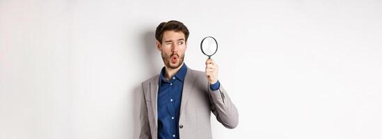 Excited funny guy in suit look through magnifying glass aside, checking out awesome thing, found something, white background photo