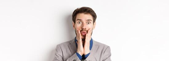 Surprised businessman in suit look with disbelied, pucker lips and touching face as staring at camera, standing on white background photo