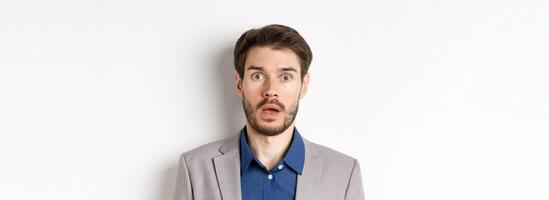 Close up portrait of shocked caucasian man in suit drop jaw, looking speechless at camera, white background photo