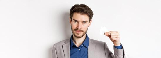 Real estate and insurance concept. Close up portrait of handsome bearded man in suit showing small paper house cutout and look assertive, white background photo