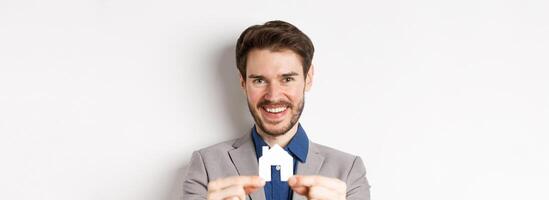Real estate and insurance concept. Handsome man in suit smiling, showing small paper house cutout, standing on white background photo