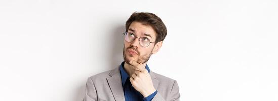 Thoughtful businessman with beard and glasses making decision, looking up and thinking, standing in suit with pensive face, white background photo