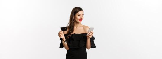 Fashion and shopping concept. Young attractive woman making purchase online, buying in internet with credit card and smartphone, white background photo