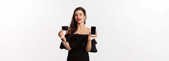 Fashion and shopping concept. Beautiful woman with red lips, winking at camera, showing smartphone screen and credit card, buying online, white background photo