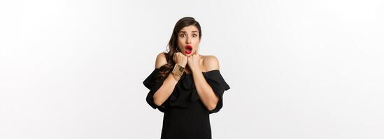 Portrait of glamour woman looking scared and shocked at camera, staring at something with fear, standing in black dress against white background photo
