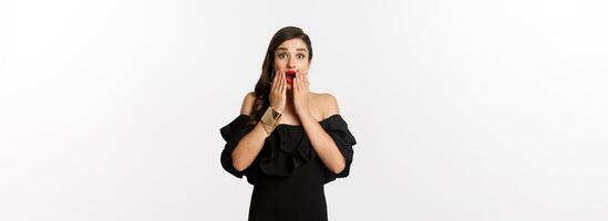 Fashion and beauty. Image of attractive female model in black dress reacting to announcement, looking amazed at camera, standing surprised over white background photo