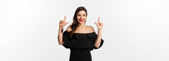 Moda y belleza. encantador mujer con rojo labios, negro vestido, sonriente contento y señalando dedos arriba, demostración logo, blanco antecedentes foto