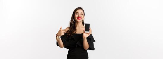 Online shopping concept. Fashionable woman in black dress pointing finger at smartphone screen, showing application, standing over white background photo