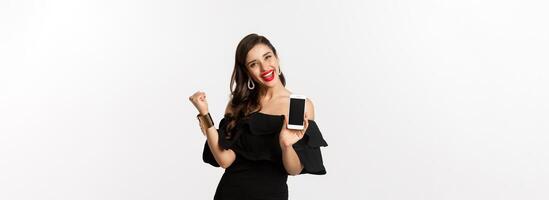Online shopping concept. Satisfied pretty woman showing smartphone screen, making fist pump to rejoice, winning in internet, standing over white background photo