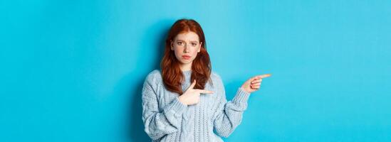 Skeptical and displeased redhead girl pointing fingers left with upset face, standing disappointed against blue background photo