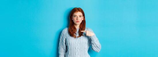 Nervous and confused redhead girl pointing at herself, standing in sweater against blue background photo
