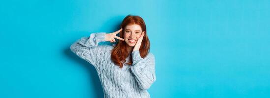 Cheerful redhead female model sending good vibes, smiling and showing peace sign, standing over blue background photo