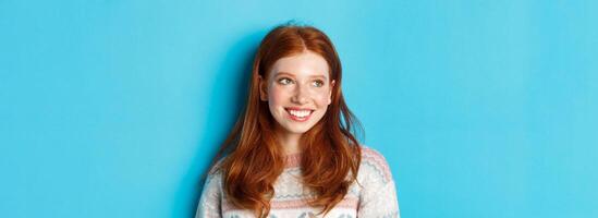 Close-up of beautiful redhead female model in winter sweater, smiling and looking left at copy space, standing over blue background photo