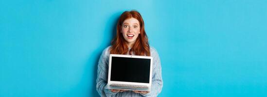Excited redhead female freelancer showing laptop screen, staring at camera amazed, standing with computer against blue background photo