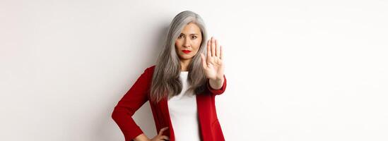 Serious asian woman in red blazer showing stop sign, telling no, frowning and rejecting something bad, standing against white background photo