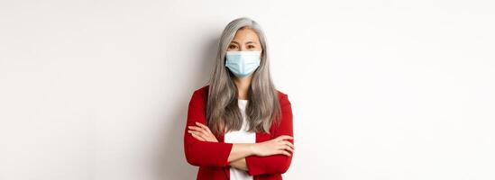 Covid-19 and business people concept. Cheerful asian lady in face mask looking at camera, cross arms on chest like professional, white background photo