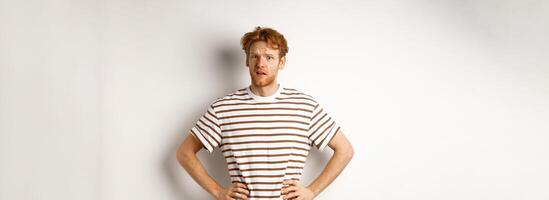 Confused and offended man with ginger curly hair staring at camera puzzled, standing questioned against white background photo