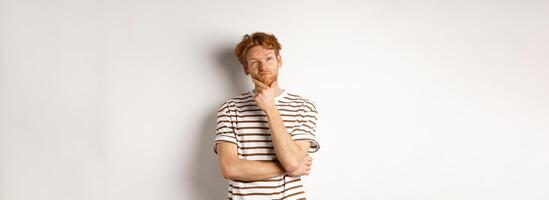 Portrait of thoughtful man with red hair thinking, looking at upper left corner and making choice, standing over white background photo
