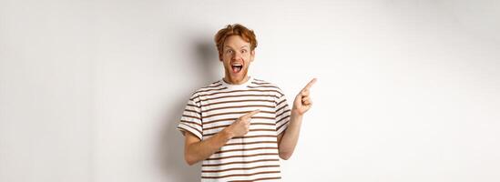 Cheerful man with red hair checking out promo offfer, pointing left at logo and smiling at camera, standing over white background photo