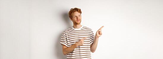 Smiling man with curly red hair, wearing striped t-shirt, smiling and pointing fingers left, demonstrate banner, standing over white background photo