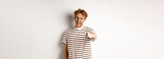 Image of funny redhead guy in glasses laughing at you, pointing finger camera and making fun of someone, standing over white background photo