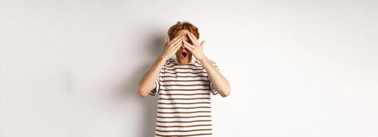 Shocked and embarrassed redhead guy covering eyes, peeking through fingers and gasping, standing over white background photo