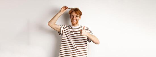 Cheerful redhead guy showing thumb-up and his red hair strand, like new color, standing over white background photo