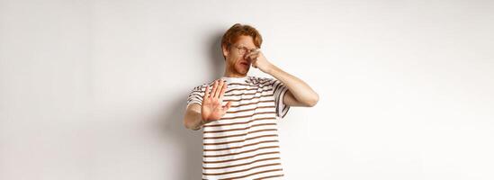Young man with red hair staring at camera and shut nose from bad smell, looking disgusted and refusing, white background photo