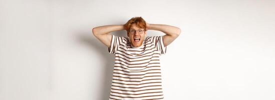 Frustrated redhead man panicking, screaming and holding hands on head, standing on white background photo