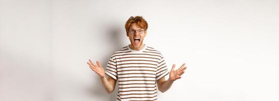 Frustrated man with red hair shaking hands and yelling at camera, looking angry and displeased, standing over white background photo