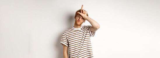Sassy and arrogant redhead man scolding lost team, showing loser gesture on forehead and making smug face, standing over white background photo