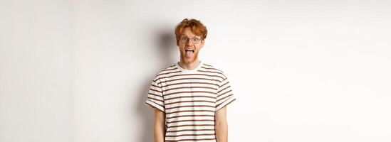 Funny young man with messy red hair and glasses showing tongue, staring at camera, standing over white background photo