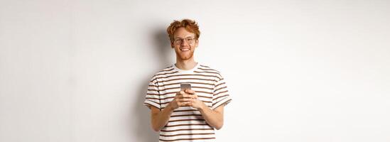 Technology and e-commerce concept. Redhead guy in glasses using mobile phone and smiling. Young man with smartphone staring happy at camera, white background photo