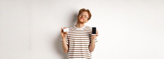 Shopping and finance concept. Pleased young man with red hair smiling from satisfaction, showing smartphone blank screen and credit card, white background photo
