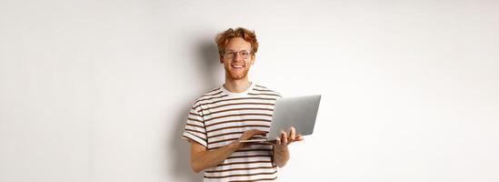 Smiling young man working on laptop and looking joyful, standing over white background photo