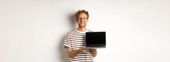 Smiling proud redhead guy showing empty laptop screen. Handsome young man demonstrating logo on computer display, white background photo